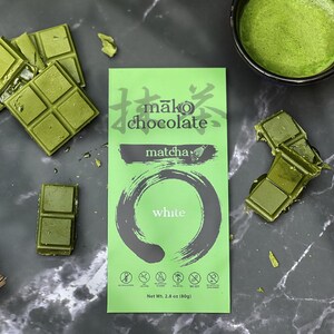 May include: A top-down shot features matcha-flavored chocolate bars and a package of Mako Chocolate Matcha White. The green chocolate is broken into squares. A small bowl of matcha tea and a bamboo whisk are also present.