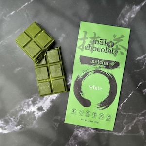 May include: Matcha-flavored chocolate bars with a bright green wrapper. The wrapper features Japanese text, the words "matcha" and "white", and a circular design. Two pieces of the green chocolate are broken off.