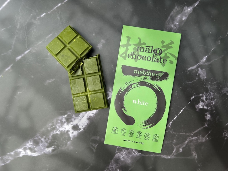 May include: Matcha-flavored chocolate bars with a bright green wrapper. The wrapper features Japanese text, the words "matcha" and "white", and a circular design. Two pieces of the green chocolate are broken off.