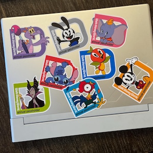 May include: A silver laptop covered in colorful stickers featuring various Disney characters. The stickers include images of Figment, Oswald the Lucky Rabbit, Dumbo, Stitch, Orange Bird, Mickey Mouse, and Maleficent, with "Walt Disney World" and "annual passholder" text.