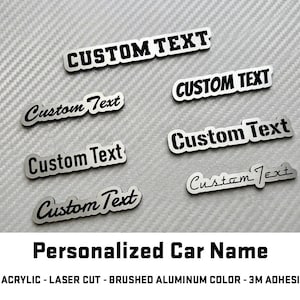 May include: A collection of custom car name badges with the text "CUSTOM TEXT" in various fonts. The badges are silver with black lettering and are made of acrylic with a brushed aluminum finish.