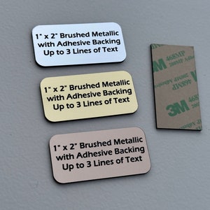 May include: Three 1 inch by 2 inch brushed metal nameplates with adhesive backing. The nameplates are white, yellow, and brown. Each nameplate has the text "1" x 2" Brushed Metallic with Adhesive Backing Up to 3 Lines of Text".
