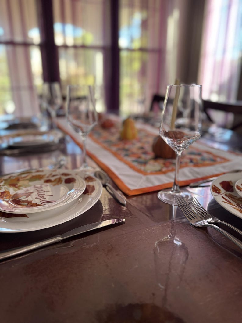 Peut inclure: Une table &agrave; manger dress&eacute;e pour un repas, avec des assiettes blanches au design automnal, des couverts et des verres &agrave; vin. Un chemin de table d&eacute;coratif avec une bordure orange et des motifs d'automne.