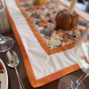 Peut inclure: Chemin de table avec un centre blanc et une bordure orange. Le chemin de table est orn&eacute; d'un motif floral color&eacute;. Une petite citrouille et une d&eacute;coration en forme de poire sont sur le chemin de table. Une cuill&egrave;re, une assiette et des verres &agrave; vin sont &eacute;galement visibles.