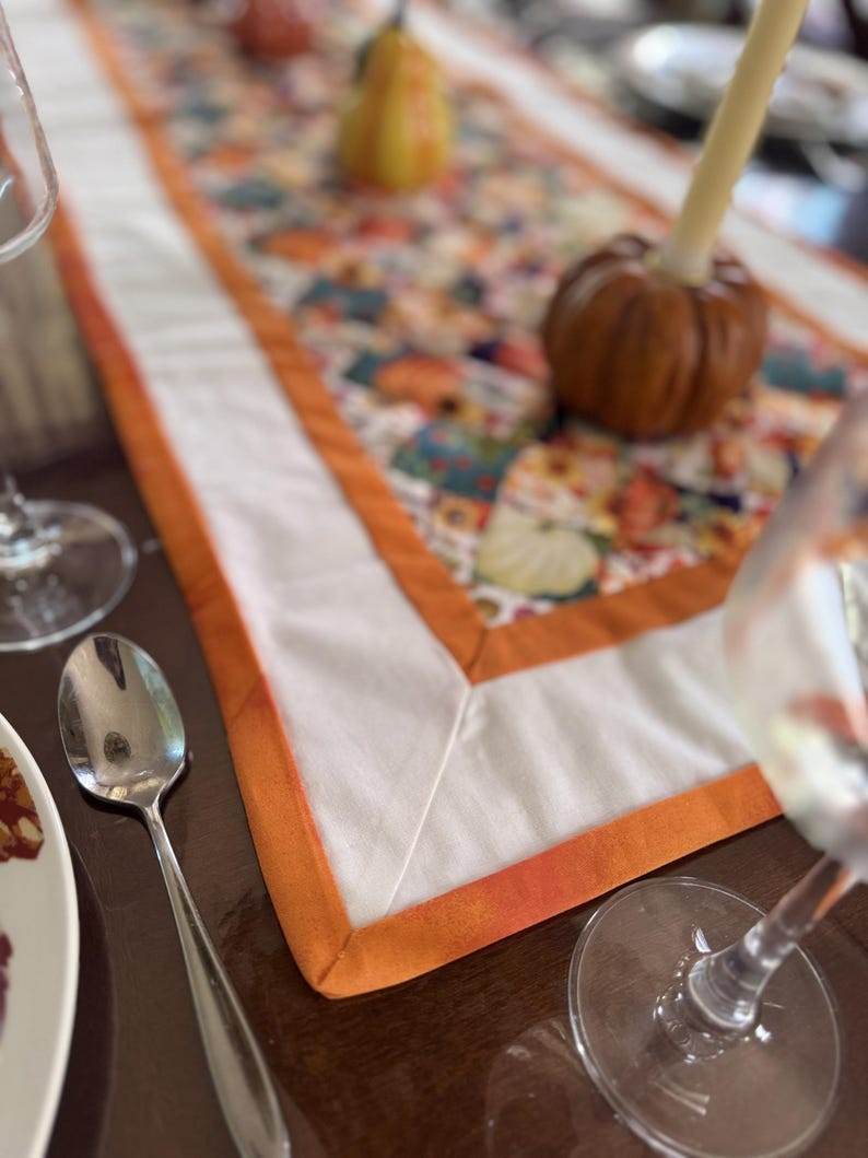 Peut inclure: Chemin de table avec un centre blanc et une bordure orange. Le chemin de table est orn&eacute; d'un motif floral color&eacute;. Une petite citrouille et une d&eacute;coration en forme de poire sont sur le chemin de table. Une cuill&egrave;re, une assiette et des verres &agrave; vin sont &eacute;galement visibles.