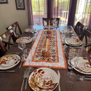 Peut inclure: Une table &agrave; manger dress&eacute;e pour un repas, avec des assiettes au motif de feuilles d'automne et le mot "Grateful". La table est orn&eacute;e d'argenterie, de verres &agrave; vin et d'un chemin de table d&eacute;coratif avec des citrouilles.
