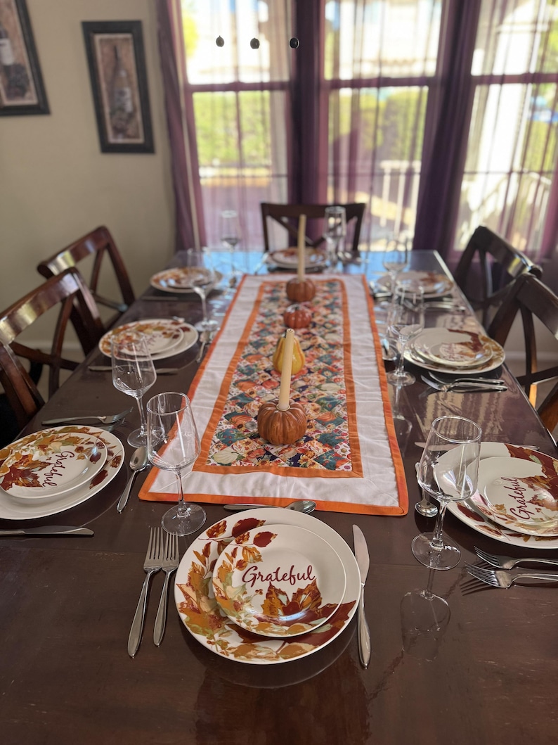 Peut inclure: Une table &agrave; manger dress&eacute;e pour un repas, avec des assiettes au motif de feuilles d'automne et le mot "Grateful". La table est orn&eacute;e d'argenterie, de verres &agrave; vin et d'un chemin de table d&eacute;coratif avec des citrouilles.