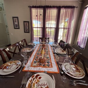 Peut inclure: Une table de salle &agrave; manger dress&eacute;e pour une occasion festive. La table est orn&eacute;e d'assiettes, de couverts et de verres &agrave; vin. Un chemin de table floral avec des citrouilles et des bougies est au centre. Les assiettes ont un motif de feuilles d'automne et le mot "Grateful".