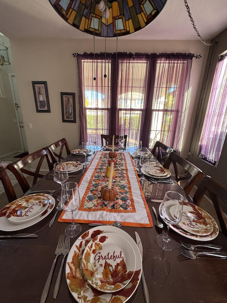 Peut inclure: Une table de salle &agrave; manger dress&eacute;e pour une occasion festive. La table est orn&eacute;e d'assiettes, de couverts et de verres &agrave; vin. Un chemin de table floral avec des citrouilles et des bougies est au centre. Les assiettes ont un motif de feuilles d'automne et le mot "Grateful".