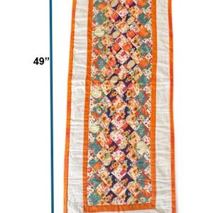 Peut inclure: Chemin de table matelass&eacute;, de 124 cm de long et 48 cm de large, avec un motif central de carr&eacute;s color&eacute;s repr&eacute;sentant des citrouilles et des fleurs. Le chemin de table a une bordure blanche avec une garniture orange.