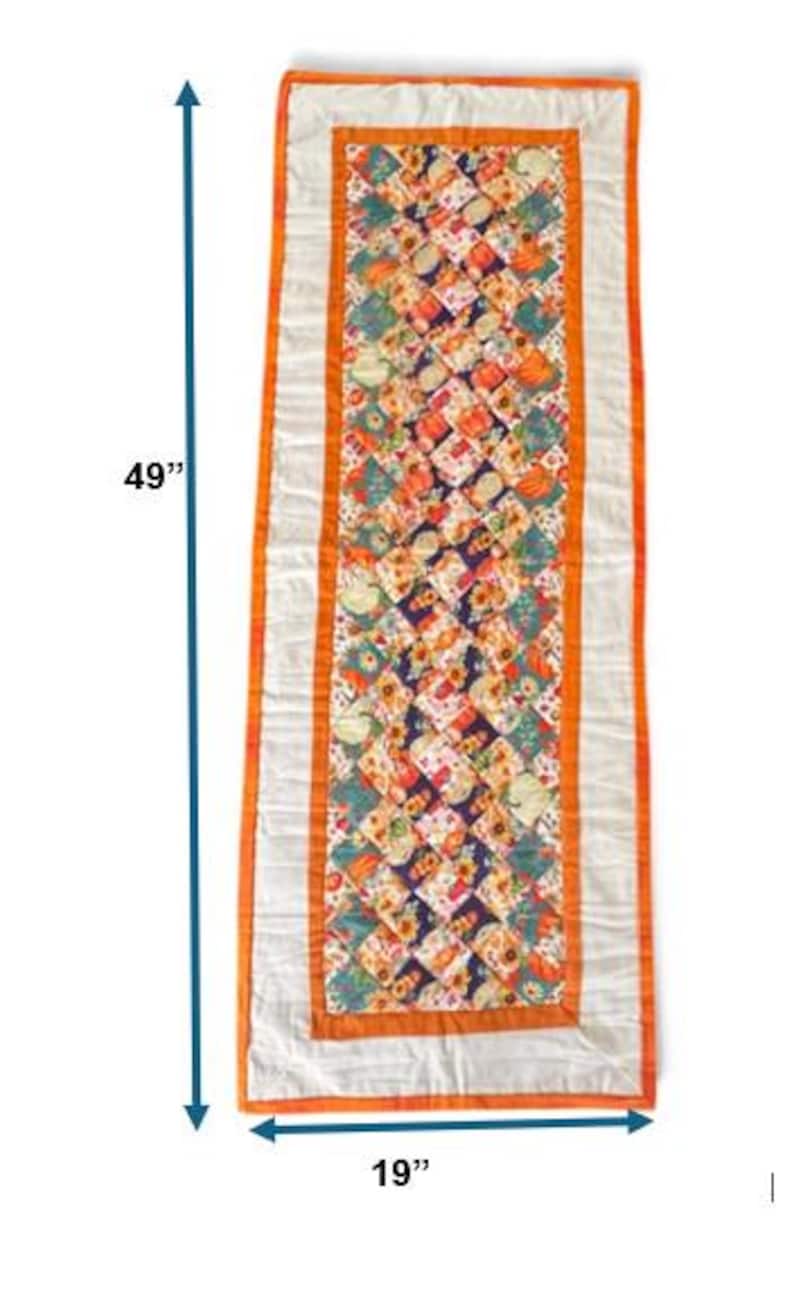 Peut inclure: Chemin de table matelass&eacute;, de 124 cm de long et 48 cm de large, avec un motif central de carr&eacute;s color&eacute;s repr&eacute;sentant des citrouilles et des fleurs. Le chemin de table a une bordure blanche avec une garniture orange.
