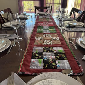 May include: A festive, quilted table runner with a patchwork design featuring Christmas-themed fabrics in red, green, and gray. The runner is placed on a dark wooden table set for a holiday meal, with plates, silverware, and wine glasses.