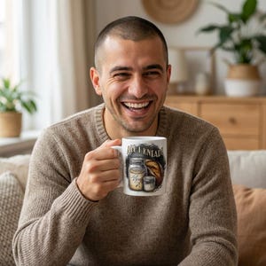 May include: White ceramic mug with the text "All I Knead!" and illustrations of bread and ingredients. The mug has a black and white design and is held by a person wearing a brown sweater.