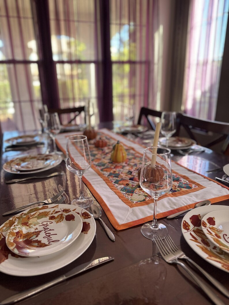 Peut inclure: Une table &agrave; manger dress&eacute;e pour un repas d'automne. Les assiettes blanches pr&eacute;sentent un motif de feuilles et le mot "Grateful". Un chemin de table color&eacute; avec des citrouilles et des courges est au centre. Verres &agrave; vin et couverts compl&egrave;tent le d&eacute;cor.