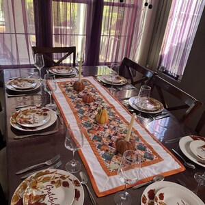 Peut inclure: Une table de salle &agrave; manger dress&eacute;e pour un repas d'automne. Les assiettes blanches pr&eacute;sentent un motif de feuilles et le mot "Grateful". Un chemin de table matelass&eacute; color&eacute; avec des citrouilles et des courges orne le centre. Verres &agrave; vin et couverts compl&egrave;tent le d&eacute;cor.