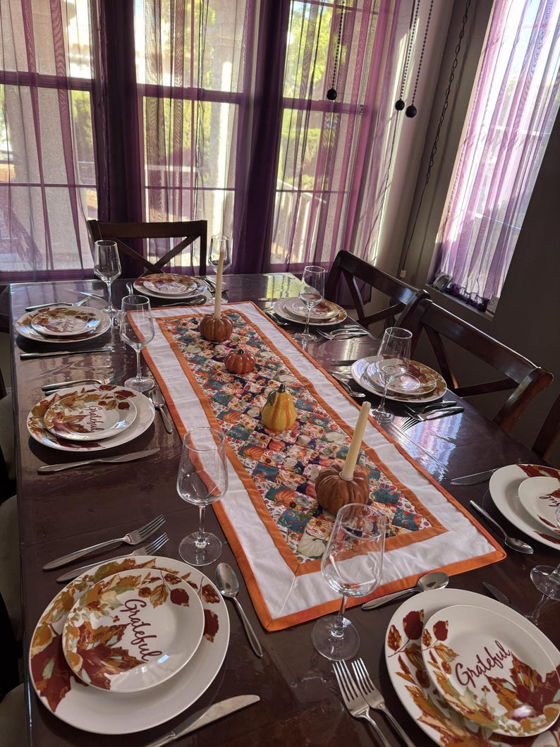 Peut inclure: Une table de salle &agrave; manger dress&eacute;e pour un repas d'automne. Les assiettes blanches pr&eacute;sentent un motif de feuilles et le mot "Grateful". Un chemin de table matelass&eacute; color&eacute; avec des citrouilles et des courges orne le centre. Verres &agrave; vin et couverts compl&egrave;tent le d&eacute;cor.