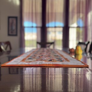 Peut inclure: Chemin de table rectangulaire avec un centre blanc et une bordure orange. Le chemin de table est orn&eacute; d'un motif floral aux tons orange, vert et jaune. Il est pos&eacute; sur une table en bois brun fonc&eacute;.