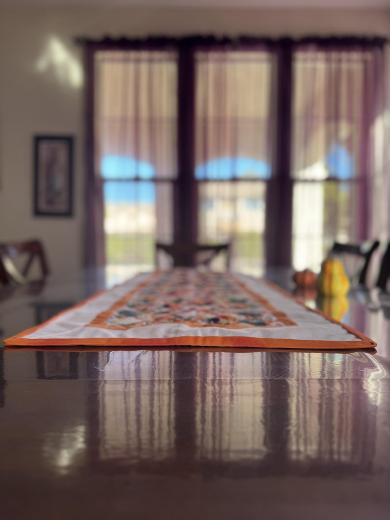 Peut inclure: Chemin de table rectangulaire avec un centre blanc et une bordure orange. Le chemin de table est orn&eacute; d'un motif floral aux tons orange, vert et jaune. Il est pos&eacute; sur une table en bois brun fonc&eacute;.