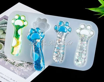 May include: Silicone mold with five paw print designs. The mold is clear and shows paw prints filled with blue, green, and silver glitter resin.