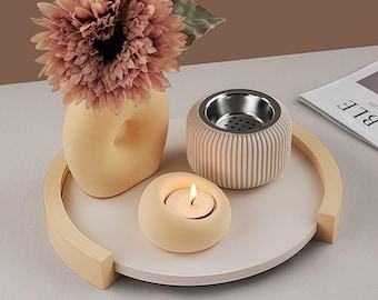 May include: A decorative beige tray holds a vase with a large pink flower, a ribbed beige container with a silver interior, and a small candle holder with a lit candle. The tray has a curved handle.