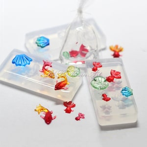 Può includere: Un set di stampi in silicone trasparente per la colata di resina, con una varietà di forme tra cui pesci, conchiglie e ninfee. Gli stampi sono mostrati con getti di resina colorati all'interno.