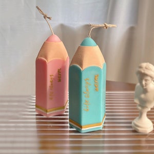 Può includere: Due candele a forma di matita, una rosa e una verde acqua, con punte beige e dettagli dorati. Ogni candela riporta la scritta "life always warm" in oro. Un cordino beige è attaccato alla sommità di ogni candela.