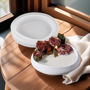 Puede incluir: Portavelas de cerámica blanca con un cuenco redondo y poco profundo. El cuenco está lleno de una vela blanca y decorado con flores secas rosas y rojas. El portavelas está sobre una mesa de madera.