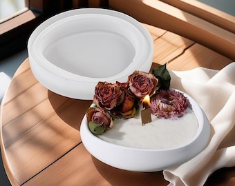 Puede incluir: Portavelas de cerámica blanca con un cuenco redondo y poco profundo. El cuenco está lleno de una vela blanca y decorado con flores secas rosas y rojas. El portavelas está sobre una mesa de madera.
