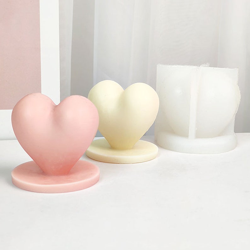 Op de afbeelding: Twee hartvormige kaarsen, &eacute;&eacute;n roze en &eacute;&eacute;n cr&egrave;me, op ronde bases. Een witte siliconen mal voor het maken van hartvormige kaarsen is ook te zien.