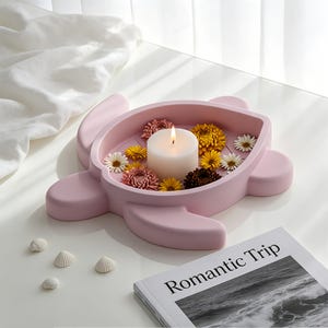 May include: A light pink turtle-shaped tray containing a white pillar candle and various small yellow, white, and dark-coloured flowers. The tray has a smooth, matte finish and is positioned near a book with the text Romantic Trip on the cover.