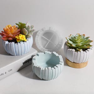 Puede incluir: Tres macetas de hormigón azul claro con un diseño a rayas. Las macetas están vacías, pero una tiene un molde de silicona para hacer las macetas.