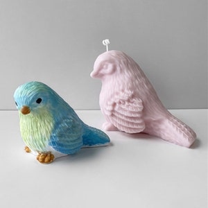 May include: Two bird-shaped objects. One is a blue and green ceramic bird with a yellow beak and brown feet. The other is a pink bird-shaped candle with a wick. Both are on a white surface.