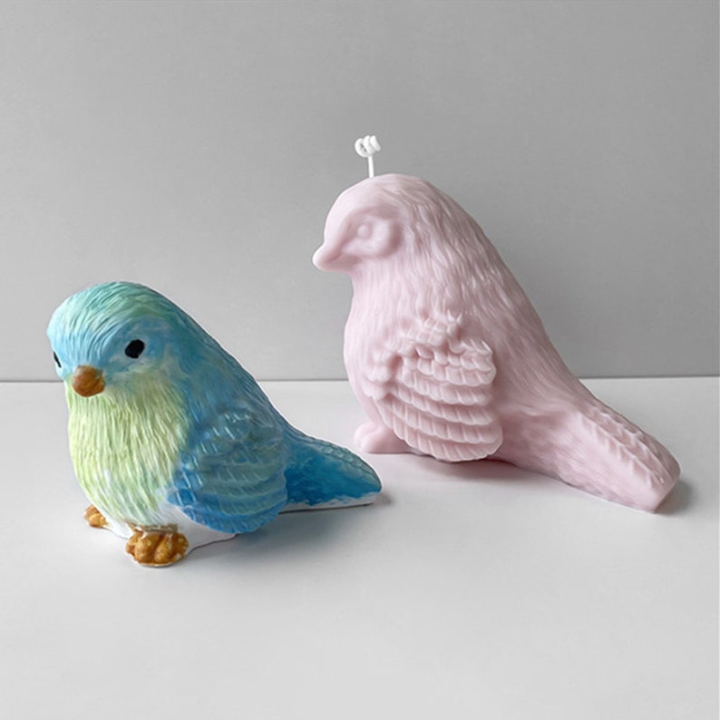 May include: Two bird-shaped objects. One is a blue and green ceramic bird with a yellow beak and brown feet. The other is a pink bird-shaped candle with a wick. Both are on a white surface.