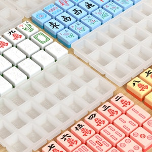 May include: A set of white, blue and pink mahjong tiles with Chinese characters and symbols. The tiles are arranged in a grid pattern on a white surface. There are also several empty tile holders.