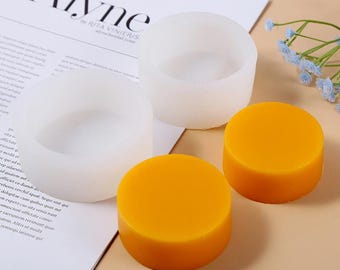 May include: Two round silicone moulds and two round yellow wax candles. The moulds are white and have a smooth surface. The candles are a bright yellow colour.