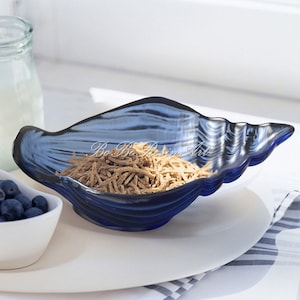 May include: A blue glass seashell-shaped bowl filled with brown cereal. The bowl is sitting on a white plate with a black and white striped napkin underneath.
