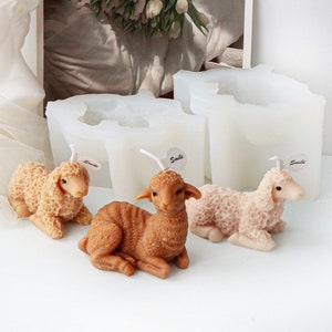 May include: Three sheep-shaped candles in brown, pink, and white, sitting on white silicone molds. Each mold has the word "Smile" on it.