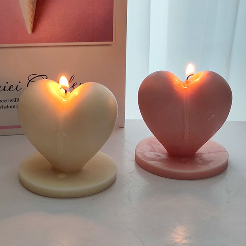 Op de afbeelding: Twee hartvormige kaarsen, &eacute;&eacute;n wit en &eacute;&eacute;n roze, zijn aangestoken en smelten. De kaarsen staan op aparte witte en roze voetjes.