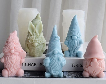Puede incluir: Cuatro moldes de jabón de colores vivos con forma de gnomo y conejo. Los moldes están hechos de silicona y están diseñados para crear esculturas de jabón detalladas. Los moldes se muestran en un libro con el título "Persiana" visible.