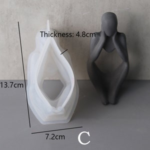 Może przedstawiać: Biała silikonowa forma do tworzenia betonowej rzeźby osoby siedzącej z głową w dłoniach. Forma ma 13,7 cm wysokości, 7,2 cm szerokości i 4,8 cm grubości. Forma jest pokazana obok gotowej betonowej rzeźby.