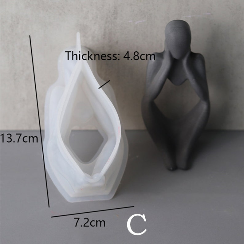 Może przedstawiać: Biała silikonowa forma do tworzenia betonowej rzeźby osoby siedzącej z głową w dłoniach. Forma ma 13,7 cm wysokości, 7,2 cm szerokości i 4,8 cm grubości. Forma jest pokazana obok gotowej betonowej rzeźby.