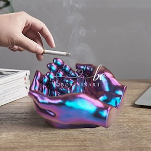 May include: A pair of hands sculpted in resin, forming a bowl shape, with a metallic iridescent finish in shades of purple and blue. The hands are cupped together, creating a space for holding cigarettes or other small items. The hands are a decorative and unique ashtray.