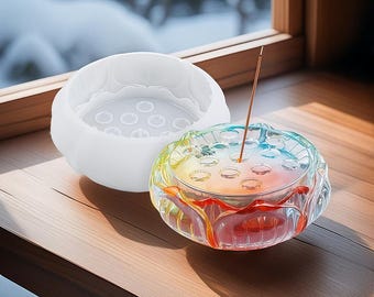 Puede incluir: Un quemador de incienso de vidrio transparente con forma de flor de loto con un degradado arcoíris. El quemador tiene varios agujeros para los bastones de incienso y está sentado sobre una superficie de madera. También se muestra un molde de silicona blanco de la misma forma.