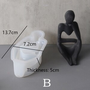 Może przedstawiać: Biała silikonowa forma w kształcie siedzącej osoby. Forma ma 13,7 cm długości, 7,2 cm szerokości i 5 cm grubości. Litera "B" jest nadrukowana pod formą.