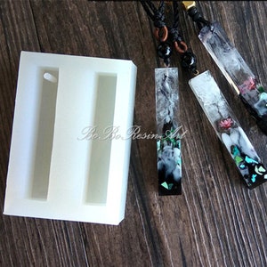 Puede incluir: Un molde de silicona blanco con dos cavidades rectangulares para hacer colgantes de resina. El molde se muestra junto a tres colgantes de resina terminados con diferentes diseños, incluyendo remolinos en blanco y negro, flores y purpurina turquesa y verde.