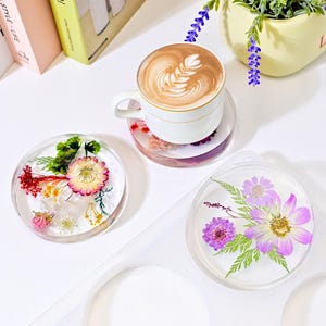 Op de afbeelding: Drie ronde, transparante onderzetters van hars met geperste bloemen erin. De onderzetters liggen op een wit oppervlak. Een onderzetter ligt onder een witte koffiekop met een latte.