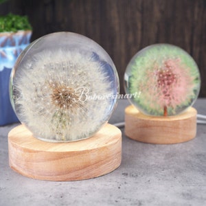 May include: Two decorative glass spheres, each containing a dandelion. The spheres sit on round wooden bases. One dandelion is white, the other is pink and green. The text "Boboresinart!" is visible on the front of the sphere.
