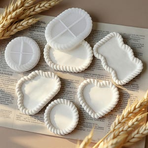 May include: A collection of white silicone molds for casting trays, featuring various shapes including oval, heart, and irregular forms. Each mold has a raised, twisted rope-style border and a smooth, flat interior surface for creating decorative items.