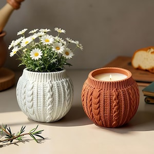 May include: Two round ceramic vessels with a textured cable knit pattern. The white vessel holds a bouquet of white daisies with yellow centers. The terracotta-colored vessel contains a lit candle with a visible white wax surface and flame.