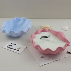 Może przedstawiać: Dwa ceramiczne świeczniki w kształcie muszli. Świecznik po lewej stronie jest jasnoniebieski, a świecznik po prawej stronie jest różowy z białą świecą w środku.